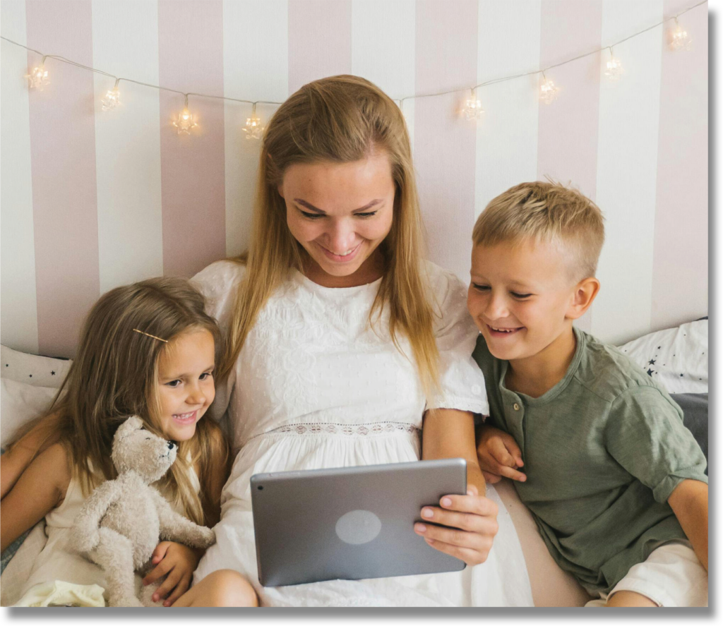 A mother and her two children smile while watching something on a tablet, sitting together on a bed with soft, pastel-colored decor.